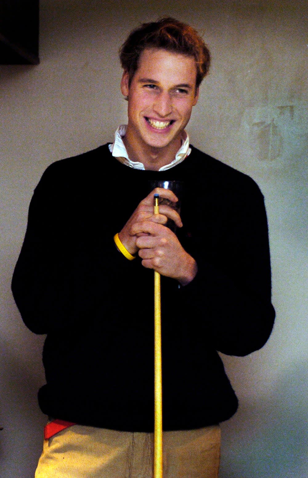 Prince William holding a billiards cue wearing a black sweater, a white collared shirt and pants.