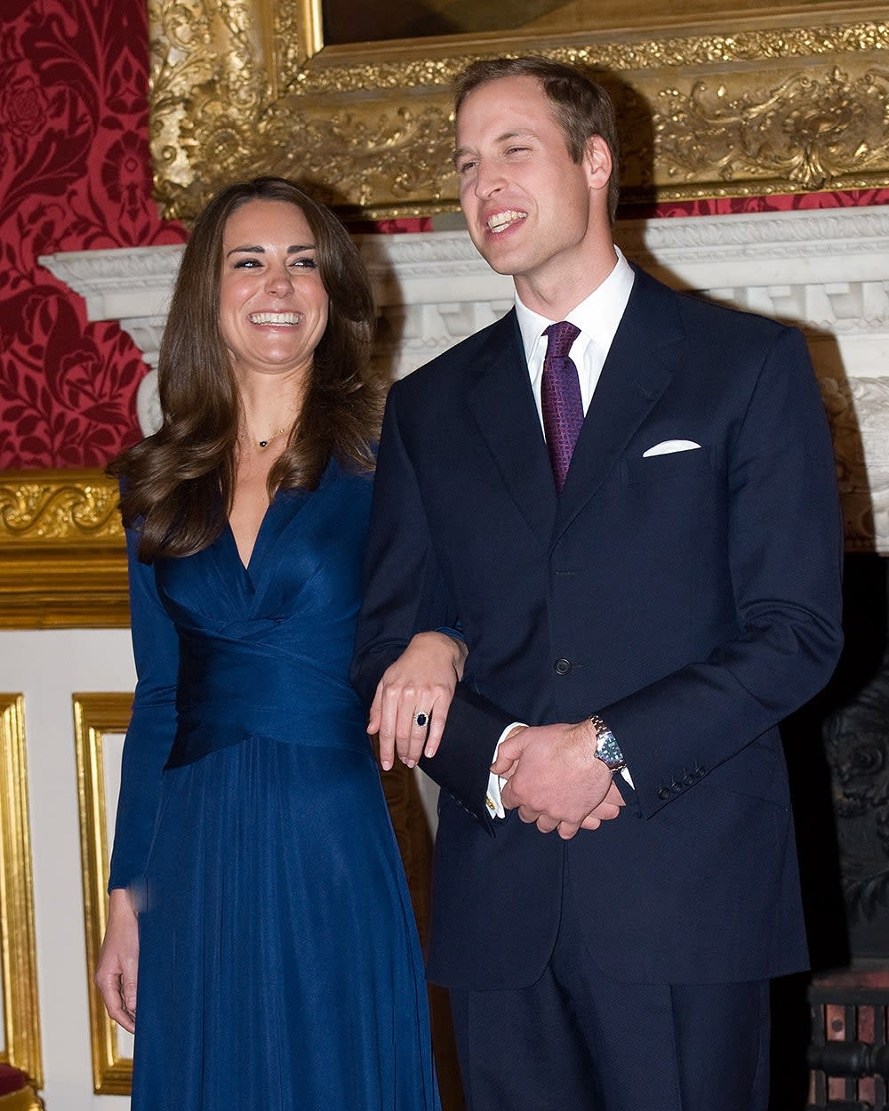 Kate Middleton smiling wearing a royal blue wrap dress in Prince William's arm. He is smiling wearing a navy suit and a purple tie.