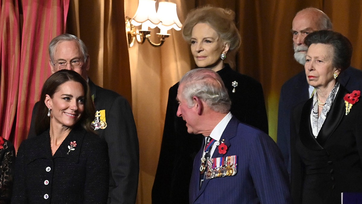 King Charles wearing a navy suit with pinned medals chatting with a smiling Kate Middleton wearing a black coat dress with pinned medals.