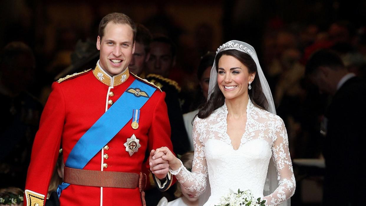 Prince William wearing a red suit with medals and a blue sash holding the hand of his smiling bride Kate Middleton