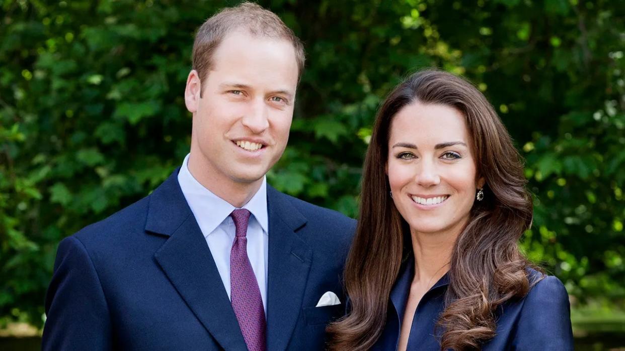 Prince William and Kate Middleton posing next to each other for a portrait wearing various shades of blue and smiling outdoors.