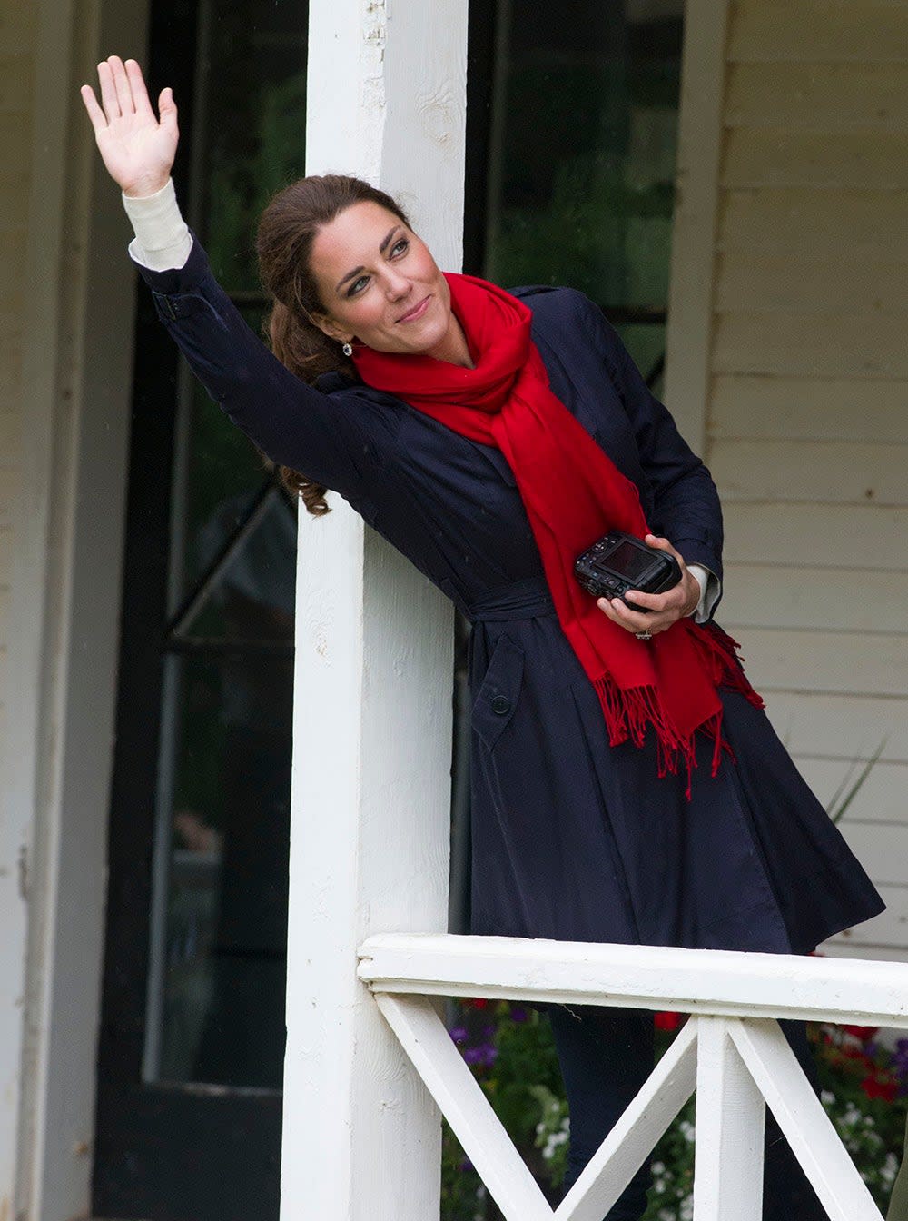 Kate Middleton holding a camera while wearing a blue coat and a red scarf waving