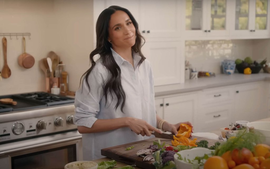 Meghan Markle standing at a kitchen counter and chopping vegetables on a cutting board