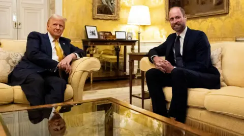 Reuters Donald Trump and Prince William seated on cream-colored sofas in a formal living room at the UK ambassador’s residence in Paris, with a glass coffee table in the foreground, ornate yellow wallpaper, and framed photographs on a table in the background.