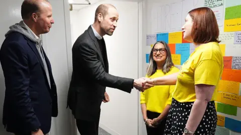 PA Media Prince William meets two women from the charity. He shakes hands with a woman wearing a yellow charity t-shirt. She is wearing a black polka dot skirt.